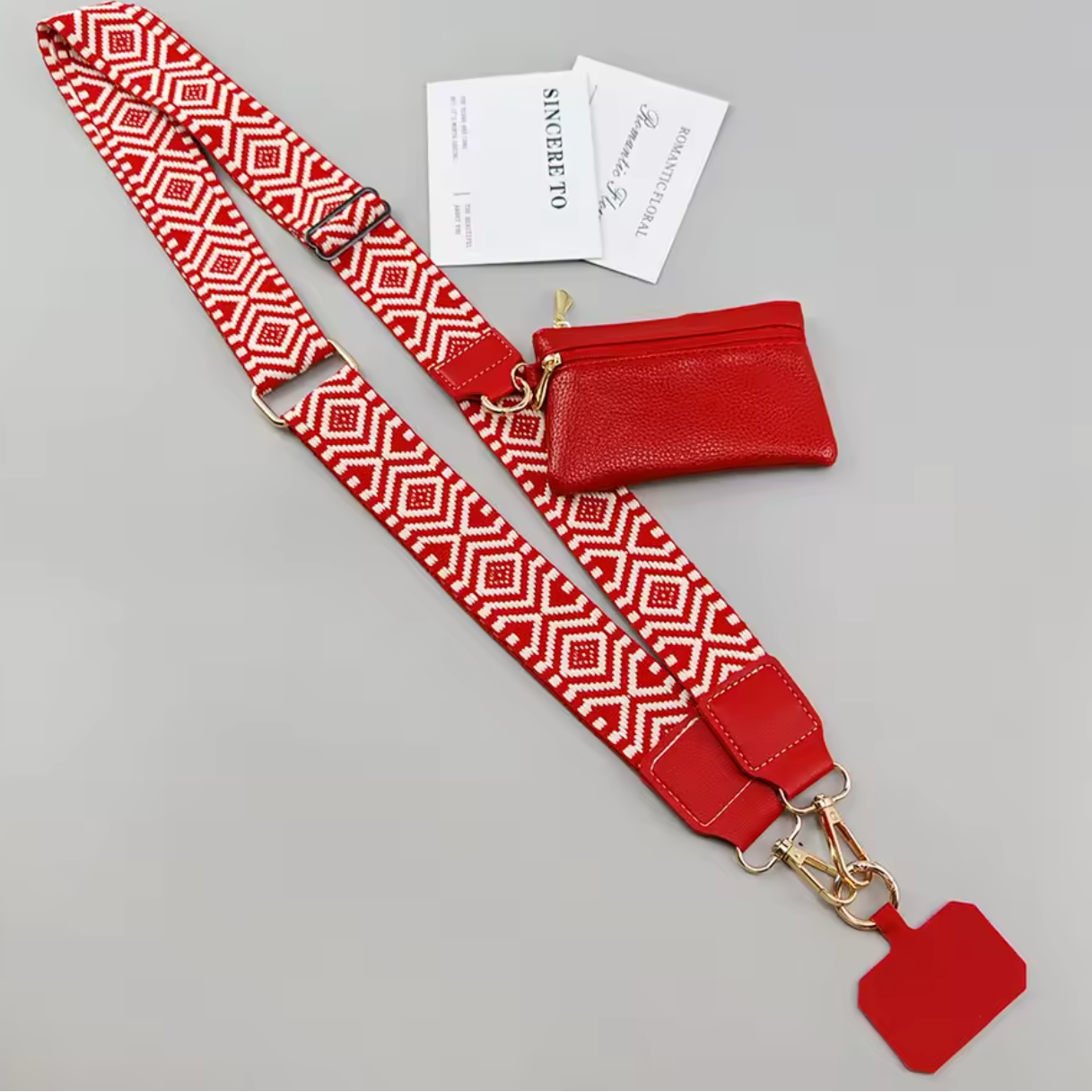 A red and white geometric patterned crossbody bag strap with gold-tone hardware, shown laid out on a light gray background alongside a matching red zip pouch, two branded cards, and a blank red tag.