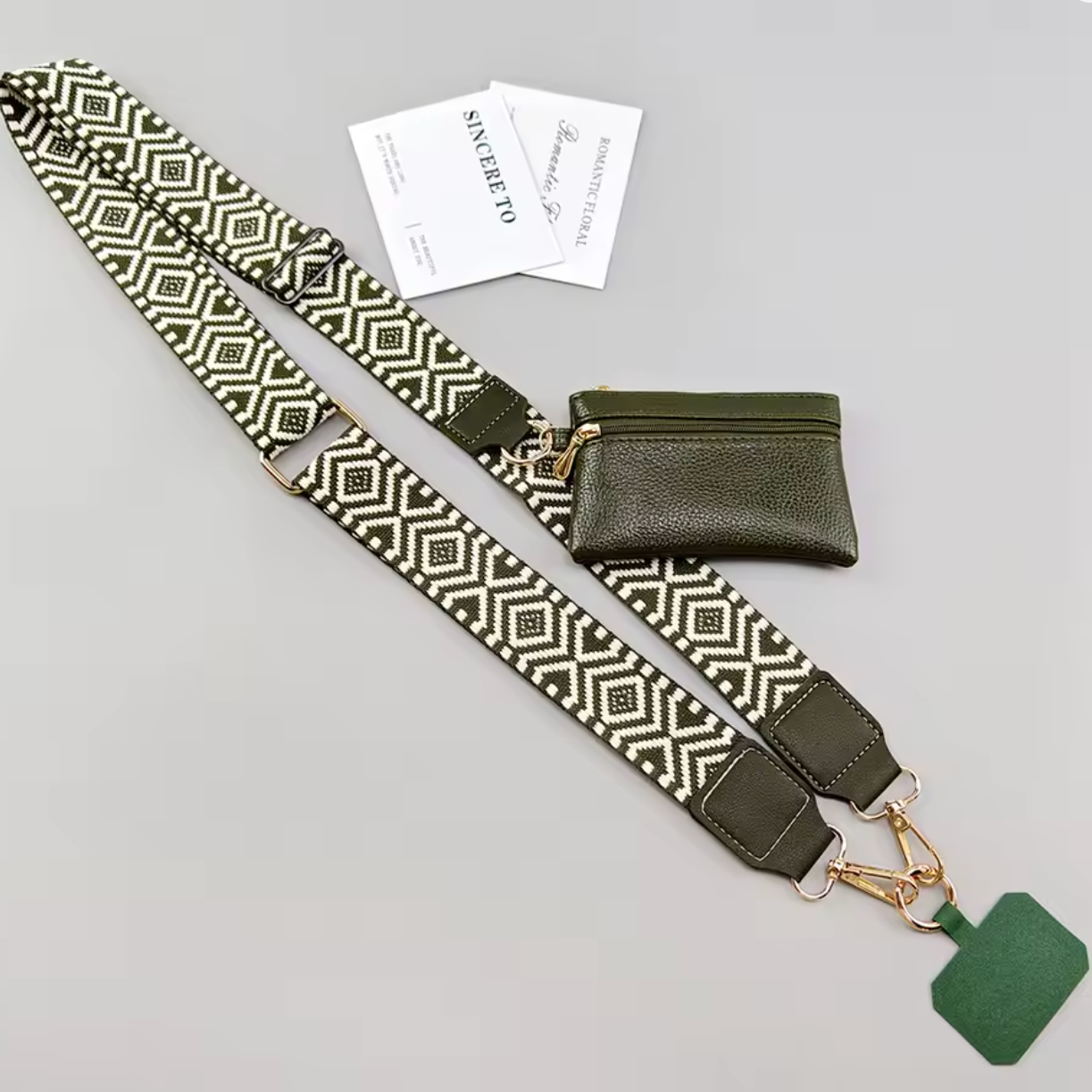 An olive green and cream geometric patterned shoulder strap with gold-tone hardware, shown laid out on a light gray background next to a matching olive green pouch and two branded cards.