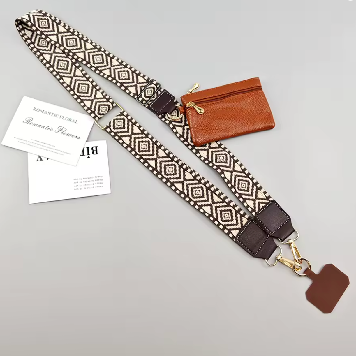 A woven shoulder strap with a geometric diamond pattern in beige and brown, featuring dark brown leather accents and gold-tone hardware. It is displayed on a light gray background alongside a small brown leather zip pouch, a white paper tag, and a card.